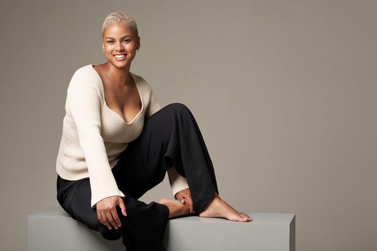 Model med kort blondt hår i beige trøje sidder på grå blok