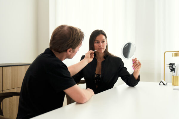 Kvinde holder spejl og ser på, mens person påfører makeup på hendes kind